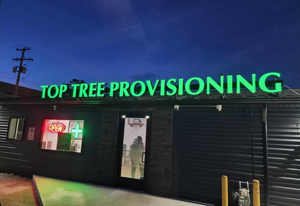 Top Tree Provisionary Center storefront at dusk with glowing green lettering in Oscoda, MI.